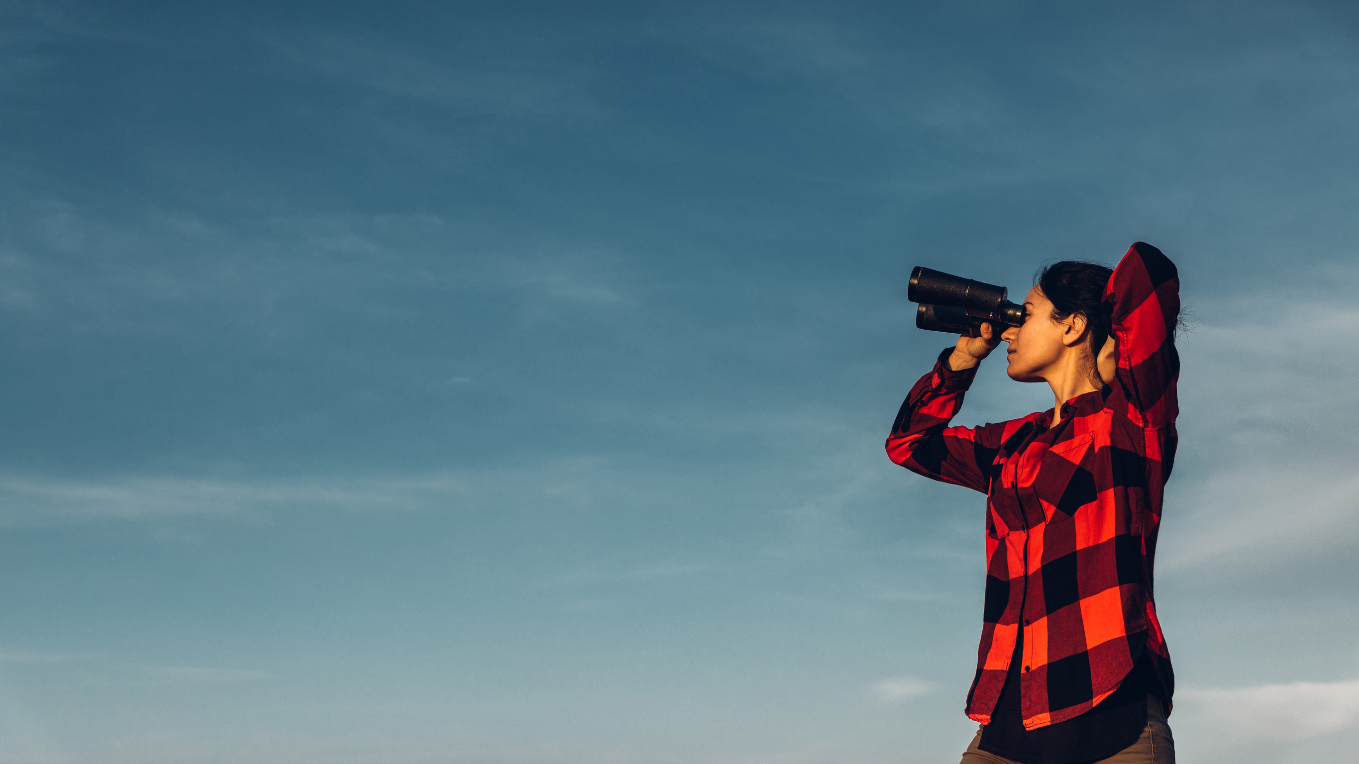 A woman wearing a red plaid shirt using binoculars to look into the distance against a clear blue sky