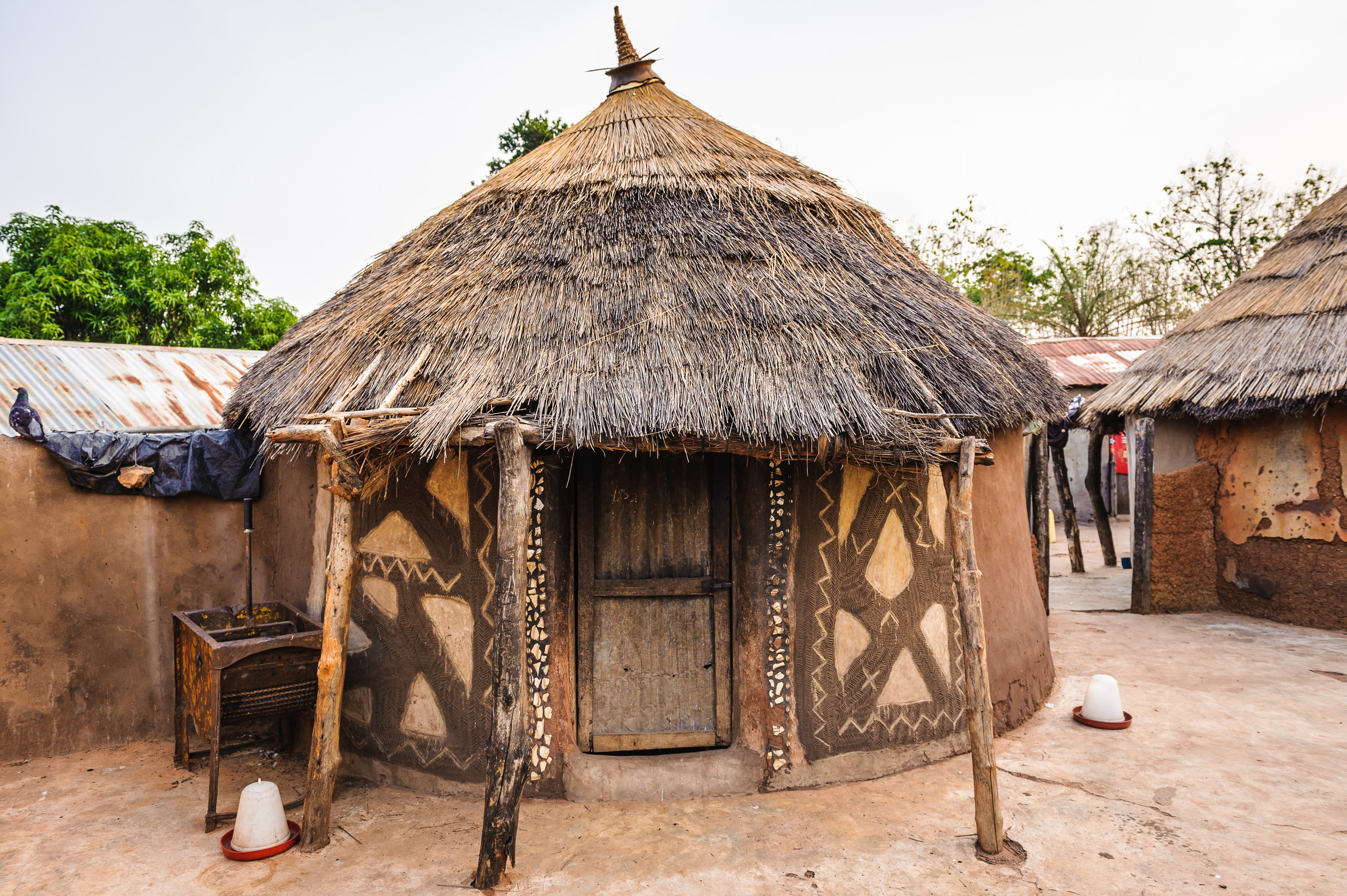 Two round huts with thatched roofs and mud walls decorated with geometric patterns. The huts are surrounded by a dirt courtyard