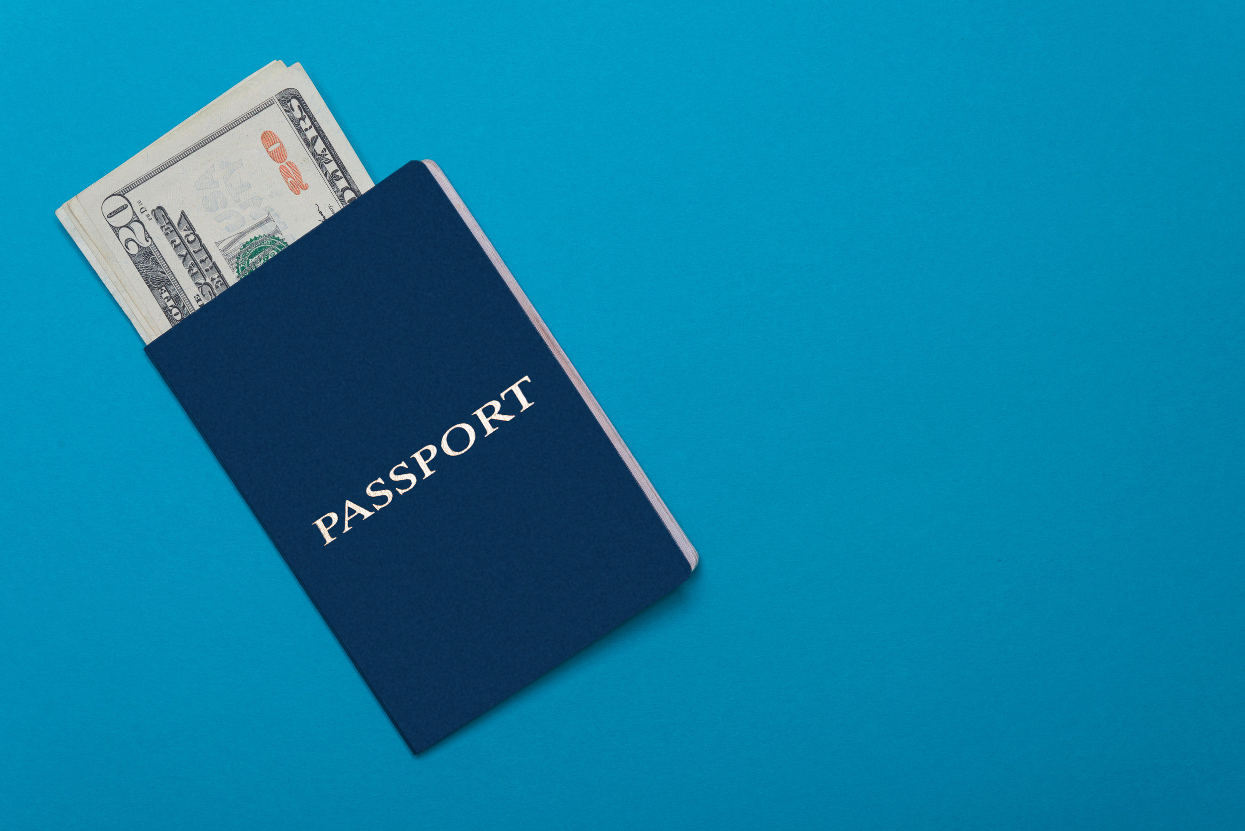 A blue passport with the word PASSPORT embossed on it, with a few US dollar bills peeking out from the side. The background is a bright blue color