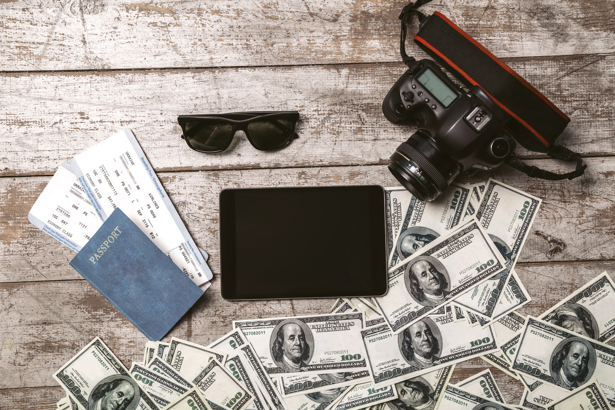 Travel essentials laid out on a wooden table, including a passport, boarding passes, sunglasses, a camera, a tablet, and US dollar bills