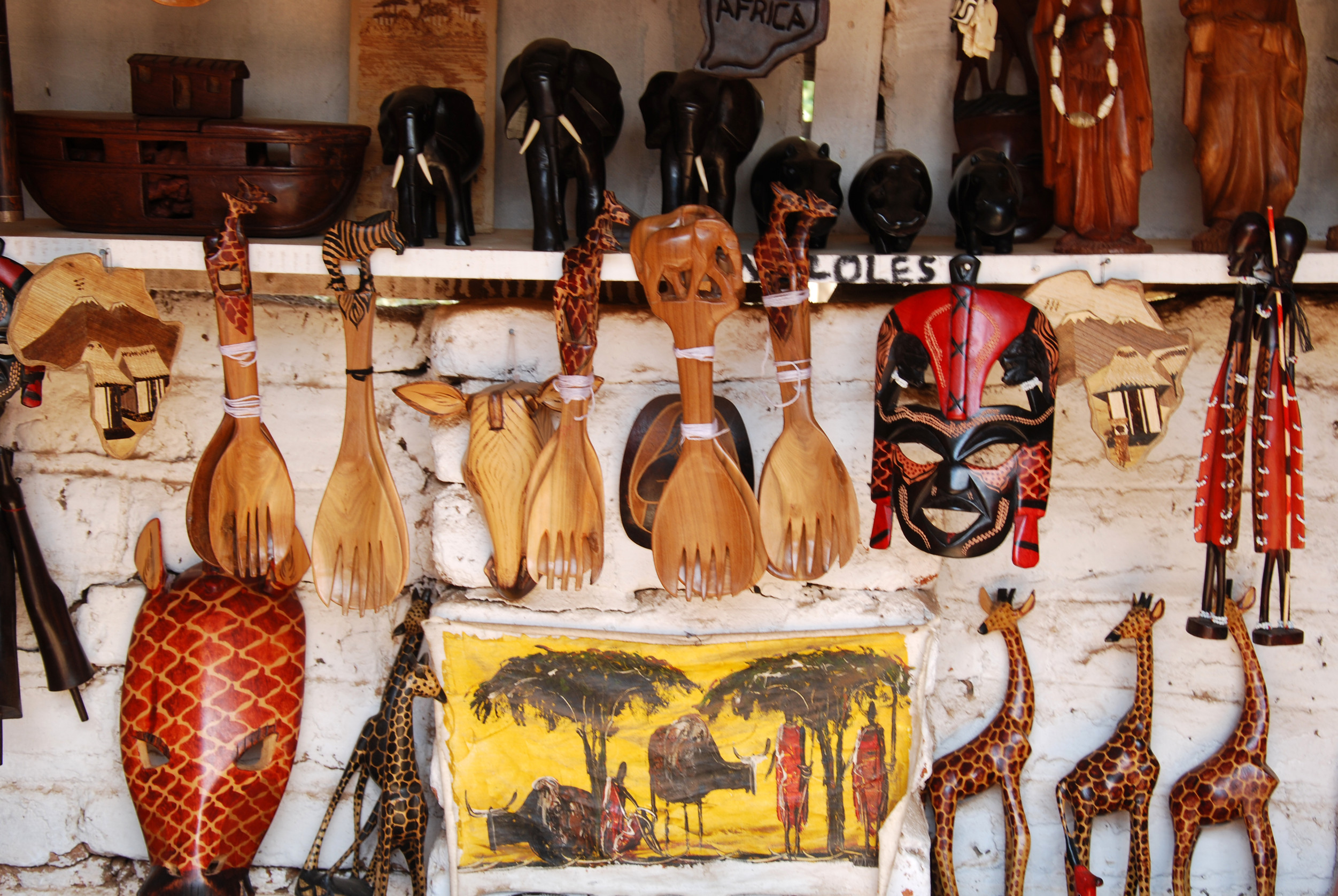 A display of colorful African handicrafts including wooden masks, figurines of animals, and utensils. The items are arranged on shelves against a brick wall