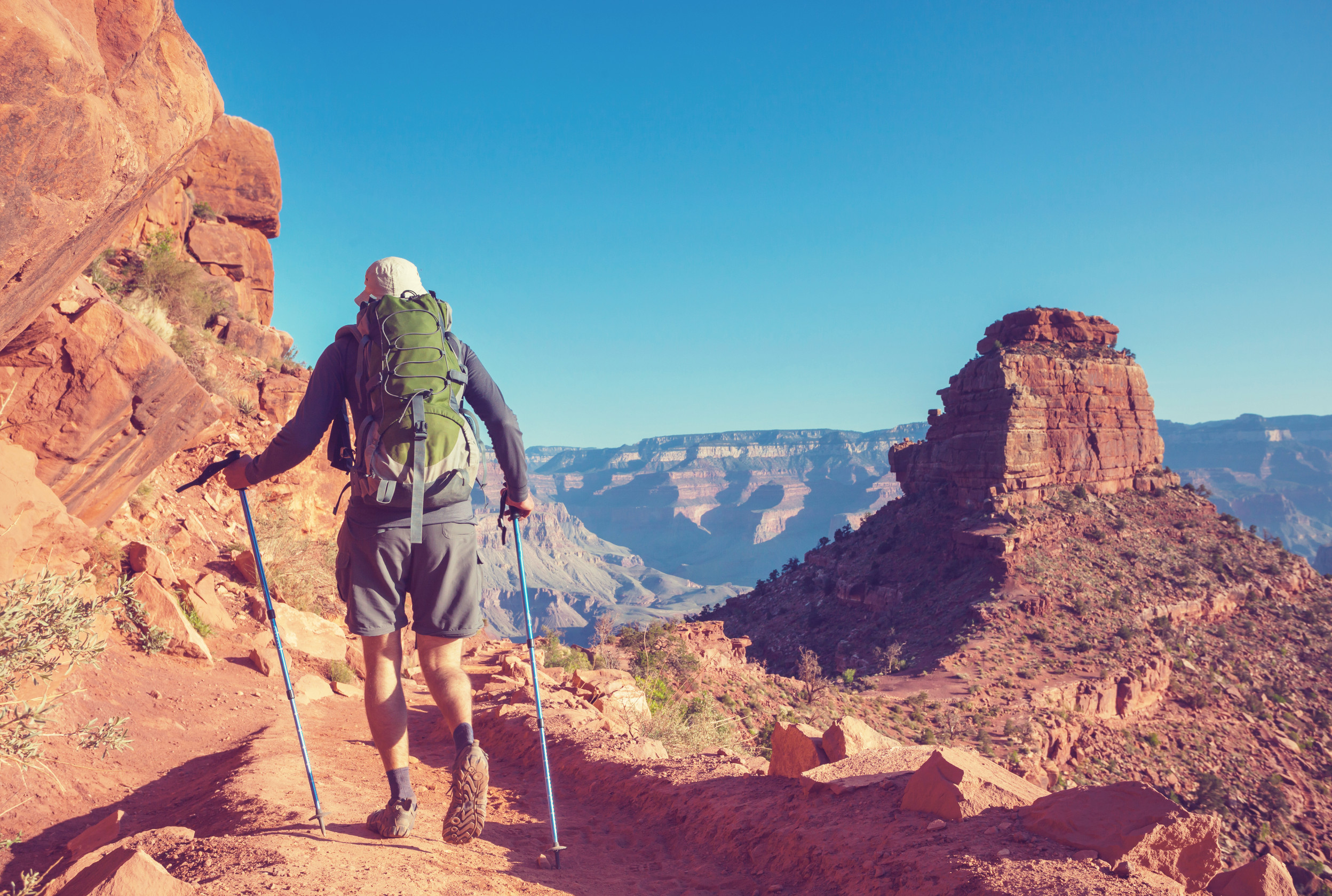 A hiker with a backpack and trekking poles walking along a rocky trail in a desert canyon