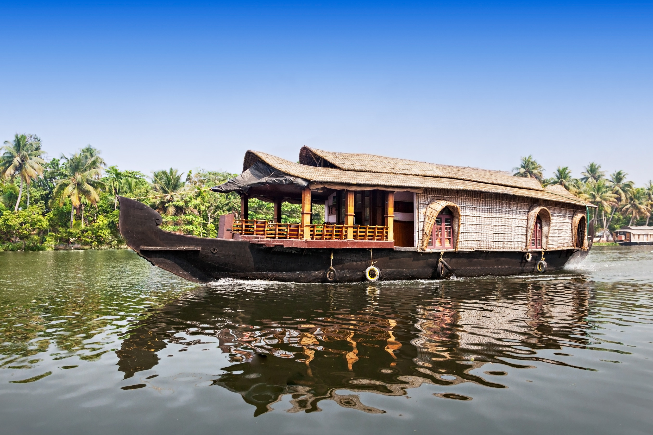 A large traditional houseboat with a thatched roof, moving through a calm river with palm trees lining the banks