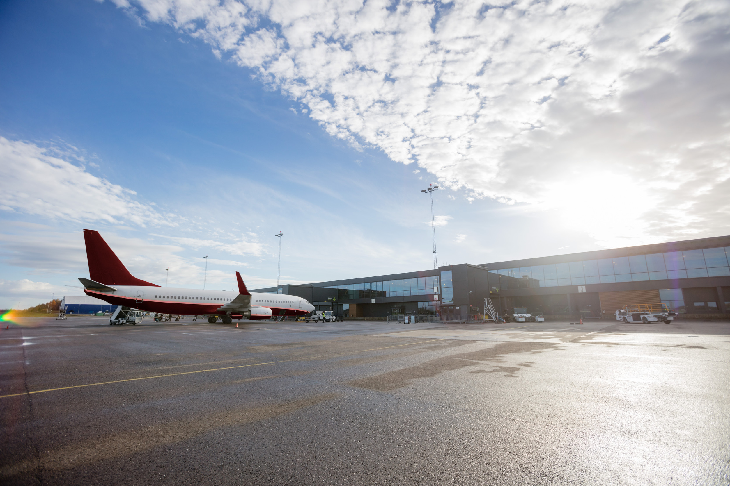 A red and white passenger jet is parked at a modern airport terminal building. The sun is shining brightly in the sky