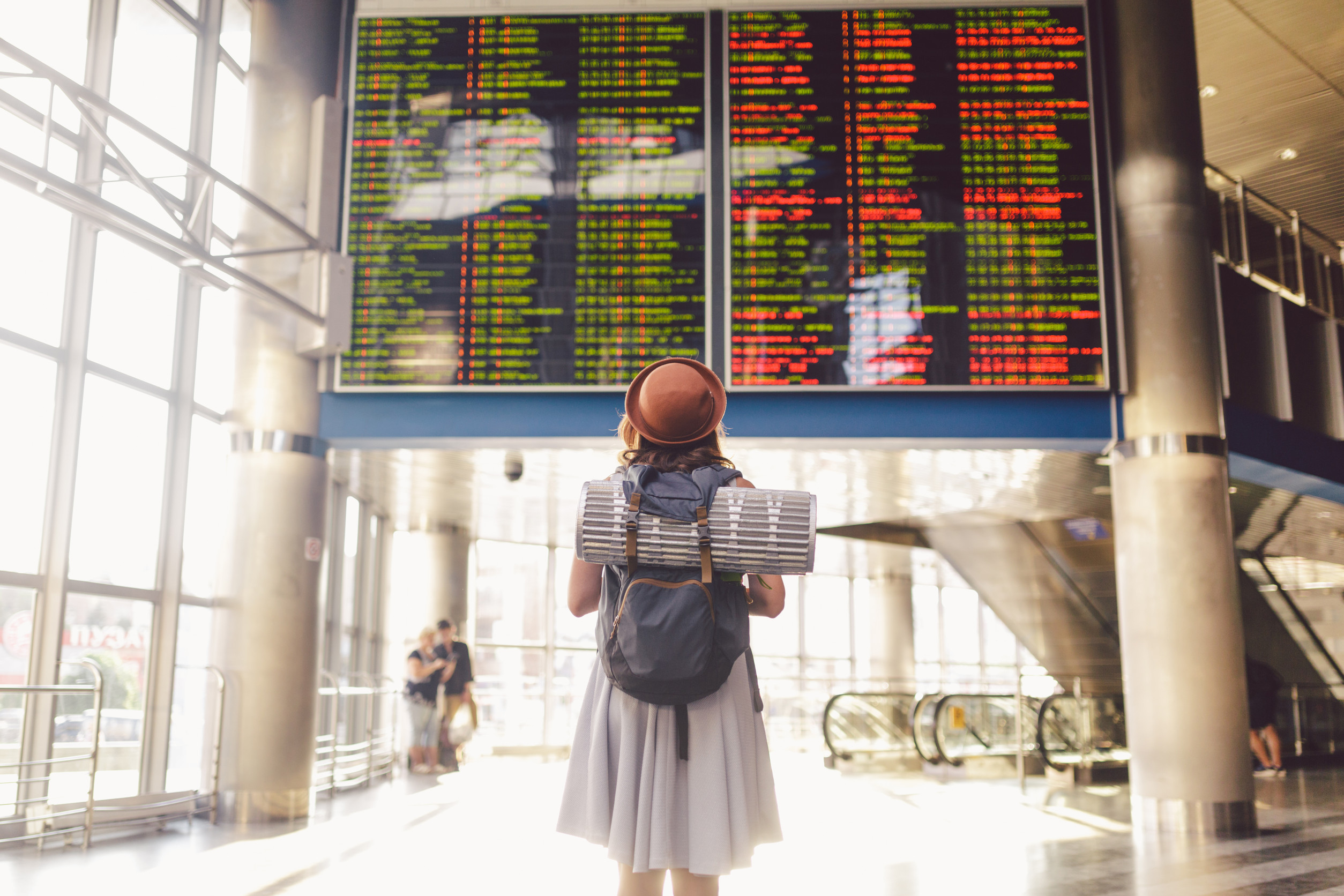 A young woman standing in front of a digital display board at an airport, which is showing flight arrival and departure information