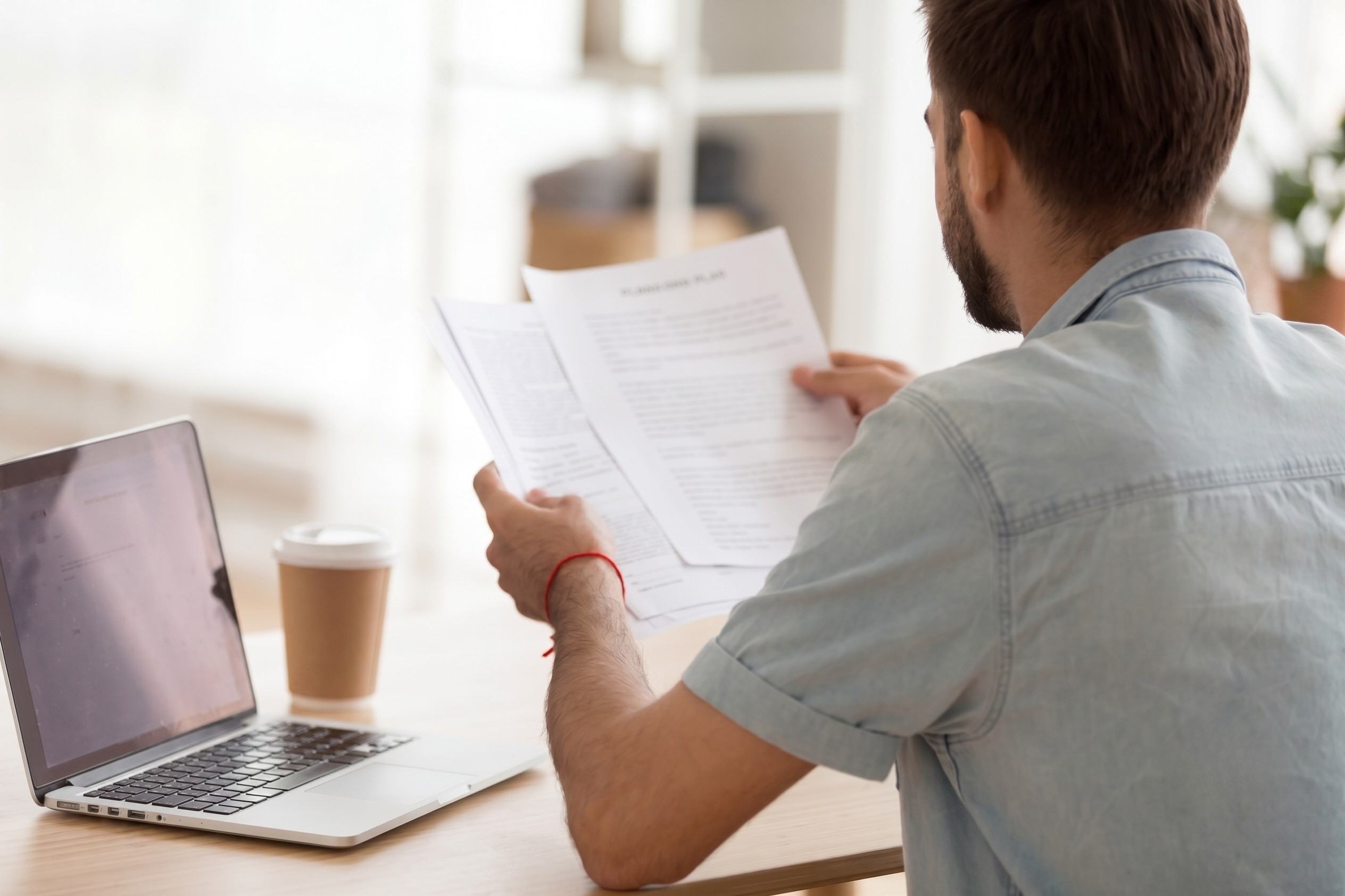 Man Looking Over Documents Next To Laptop