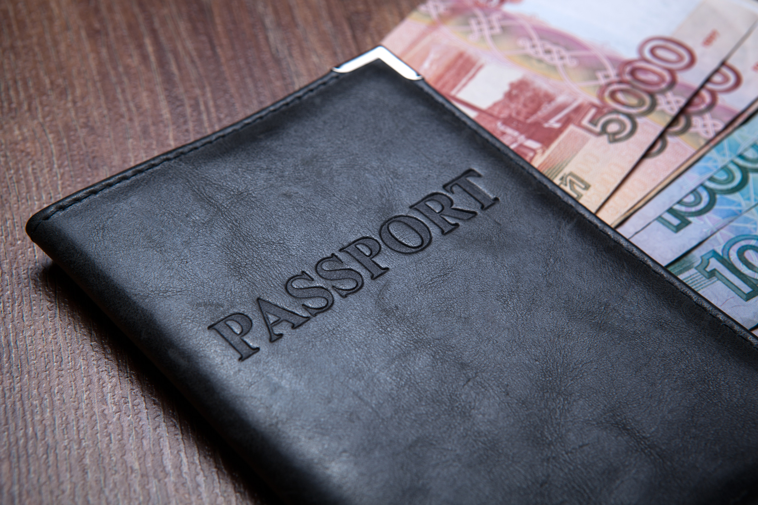 A black passport cover with the word 'PASSPORT' embossed on it, lying on a wooden surface with banknotes visible