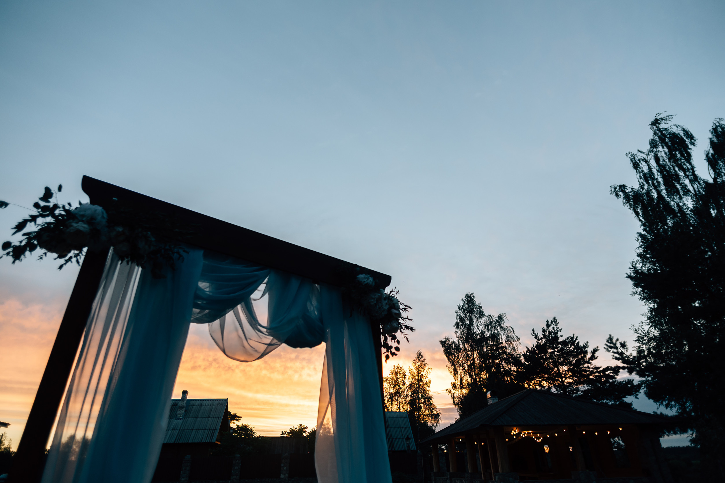 A wooden wedding archway adorned with white fabric and flowers, set against a vibrant sunset sky