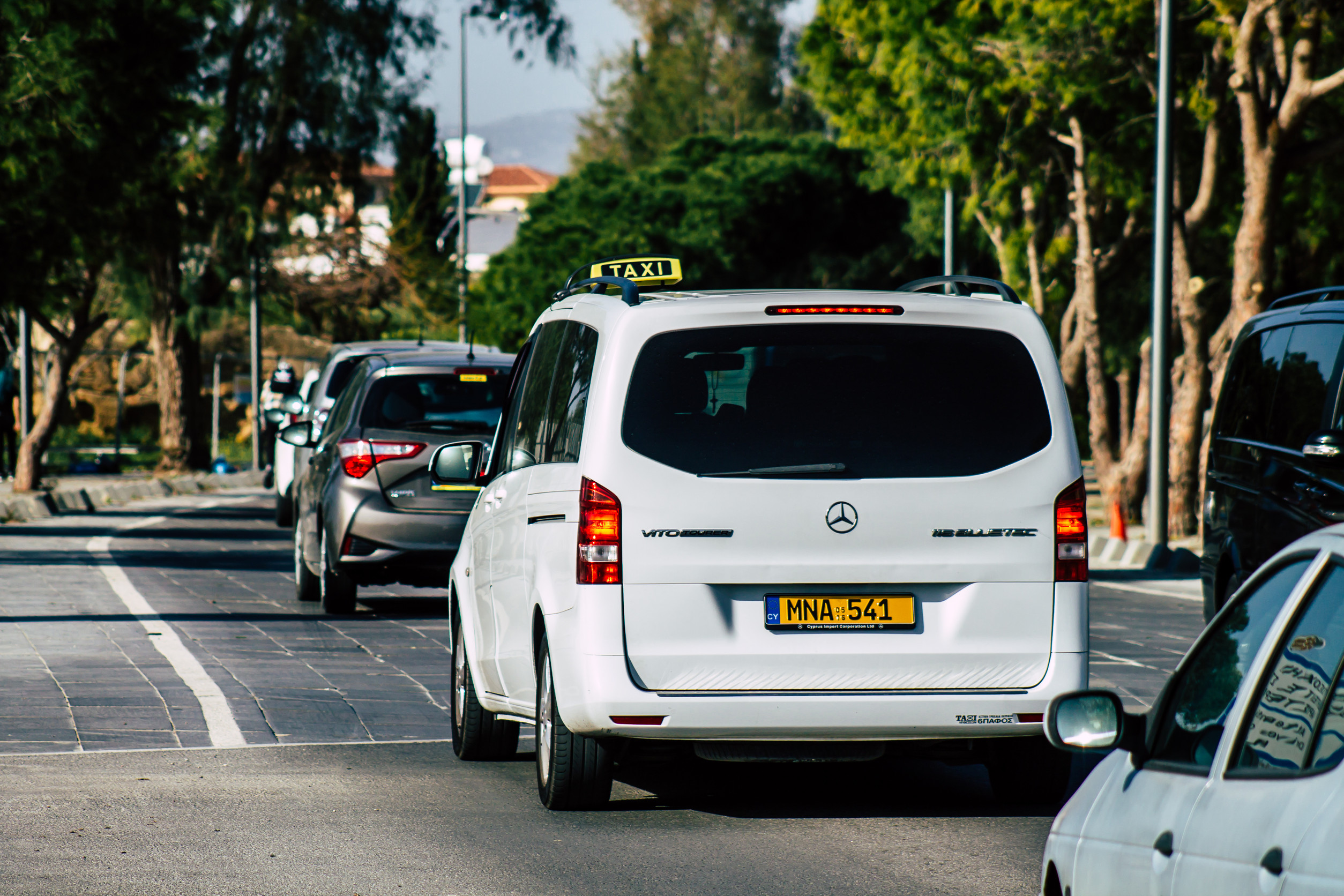 White Mercedes-Benz taxi van driving on a tree-lined stree