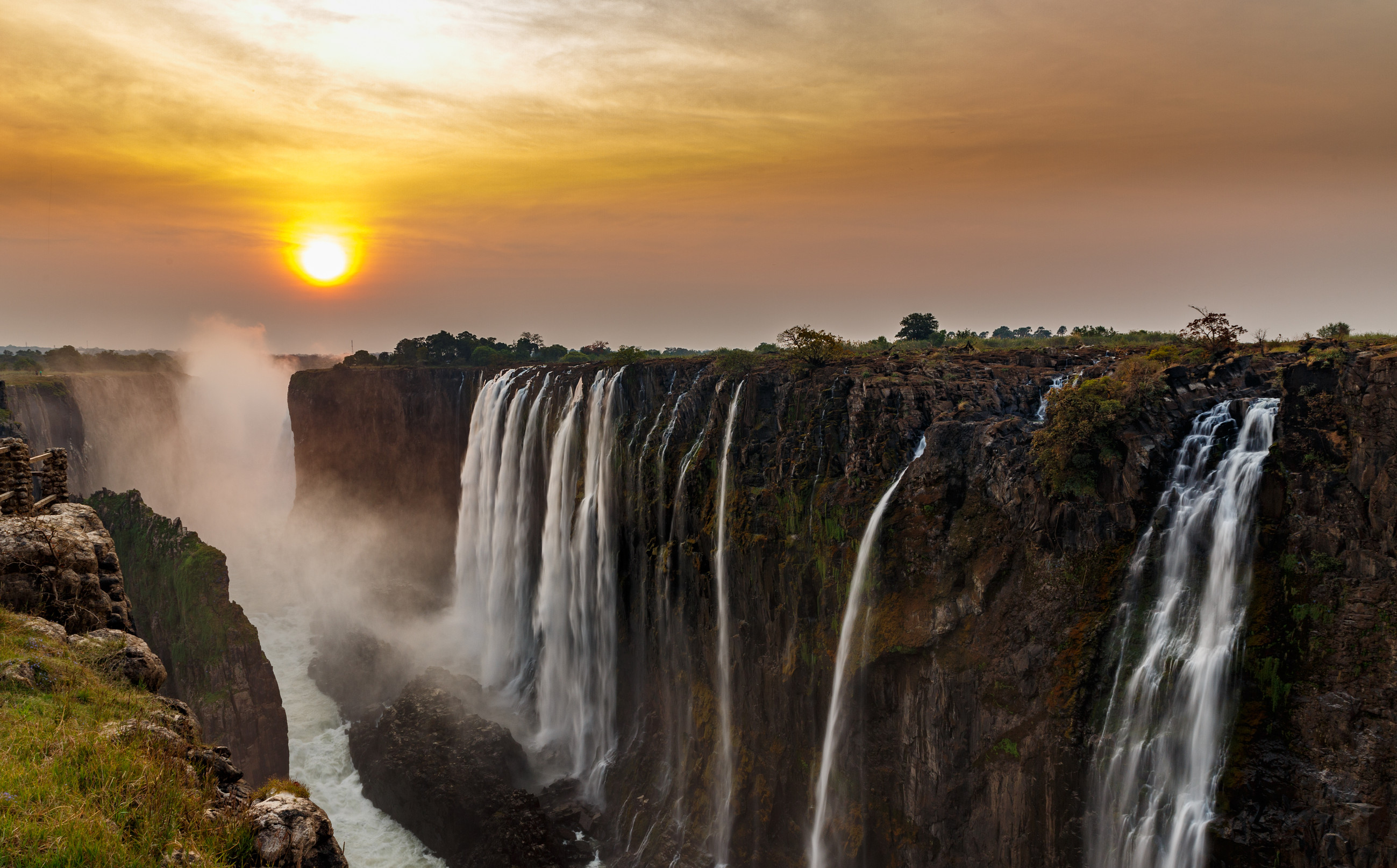 A panoramic view of Victoria Falls at sunset, with the setting sun casting a golden light on the water mist and the surrounding landscape